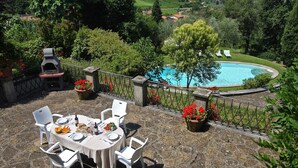 A patio with a table set for four, surrounded by stone flooring and white chairs. A stone barbecue grill and potted red flowers are visible. In the background, there is a swimming pool with a view of a lush green landscape. - Villa Lucolena - Villa with private pool (Lucolena In Chianti)