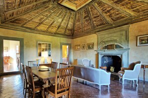Ein Zimmer mit hoher Holzbalkendecke, Kamin, Esstisch mit Stühlen und einem großen Fenster.
