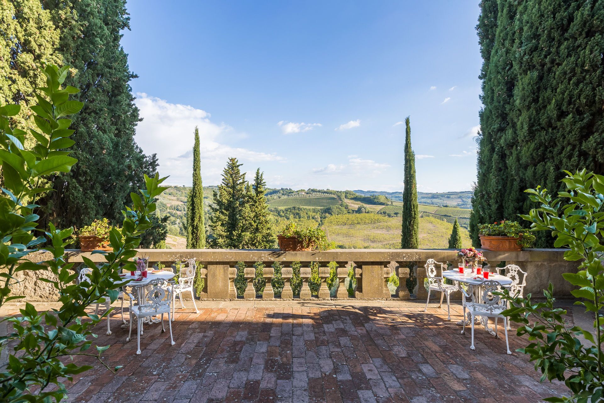 Une terrasse dotée de balustrades en pierre, de chaises et de tables en métal blanc, offrant une vue sur un vignoble et des collines ondoyantes.
