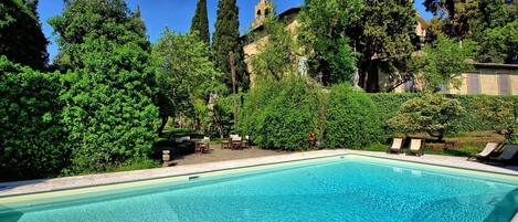 A swimming pool surrounded by lush greenery and a stone building with a tower.