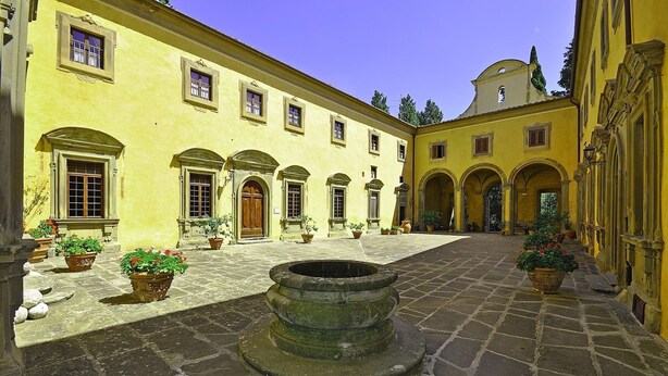 Ein Innenhof mit einem zentralen Brunnen, gelbe Gebäude mit Rundbogenfenstern und Kübelpflanzen.