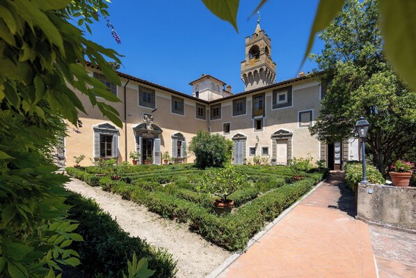 A courtyard with a building featuring a tower, surrounded by greenery and a paved walkway.