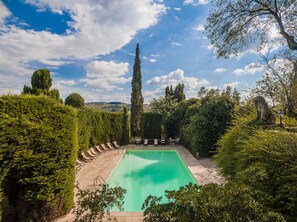 En rektangulär pool omgiven av välklippta häckar och höga träd, med en klar himmel ovanför.