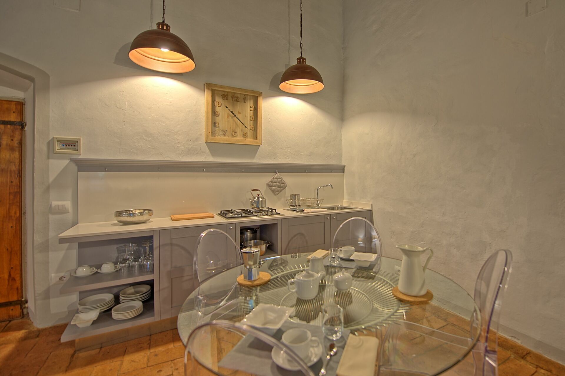 Une cuisine avec une table ronde en verre, deux lampes suspendues, une horloge en bois et un évier.