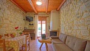 A cozy room with stone walls, a wooden ceiling, a fireplace, a sofa, a dining table with chairs, and a laptop on a wooden stool. - Villa Nipos - Villa Rental on the Island of Crete, Greece (Vrýsai)
