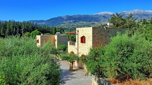 A stone building with a pathway leading to it, surrounded by greenery and mountains in the background. - Villa Nipos - Villa Rental on the Island of Crete, Greece (Vrýsai)