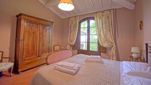 Une chambre avec une grande armoire en bois, un lit recouvert de draps blancs et une fenêtre ornée de rideaux à motifs floraux.