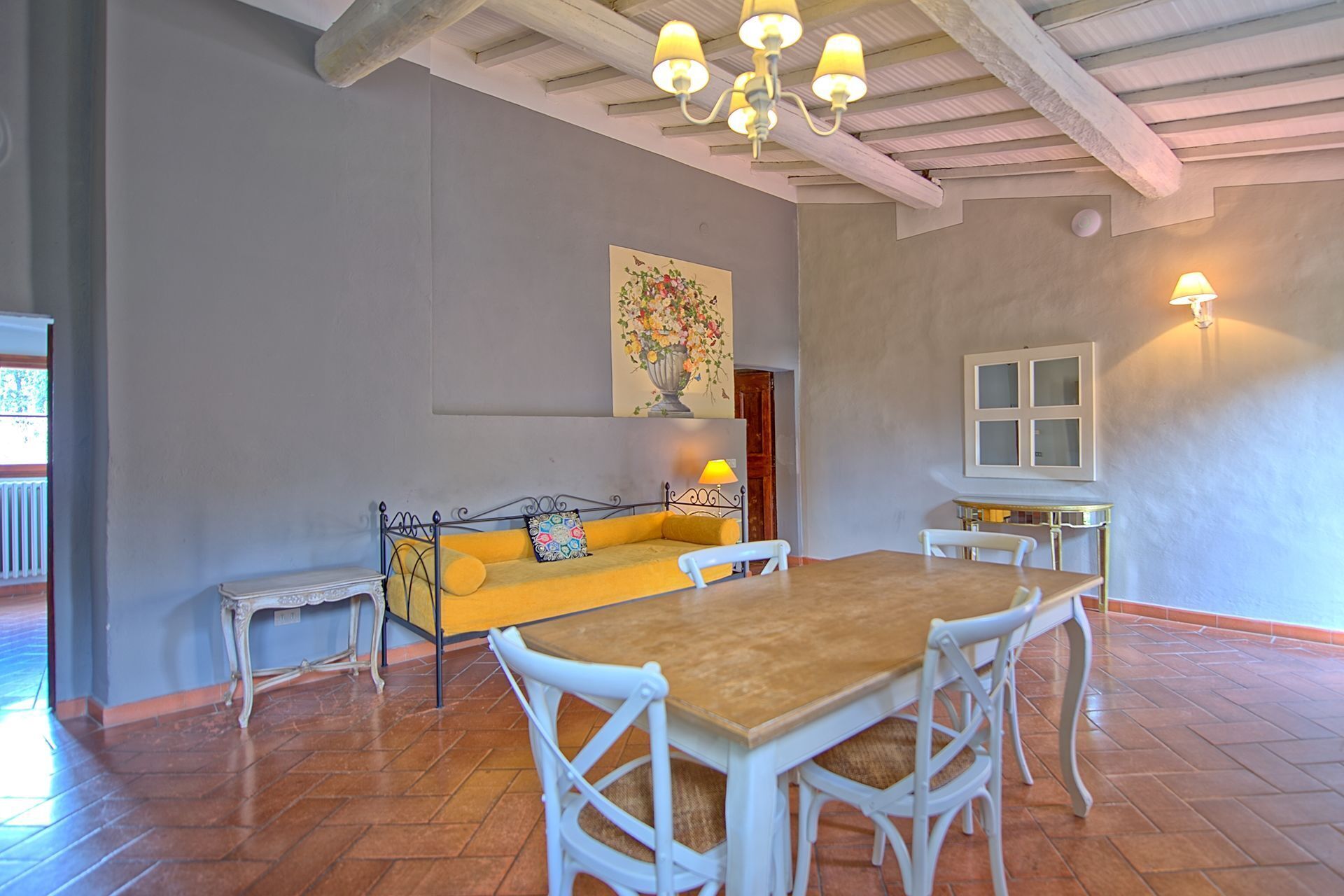 Un coin repas avec une table en bois et des chaises blanches, un canapé jaune agrémenté de coussins à motifs et un tableau représentant des fleurs accroché au mur.