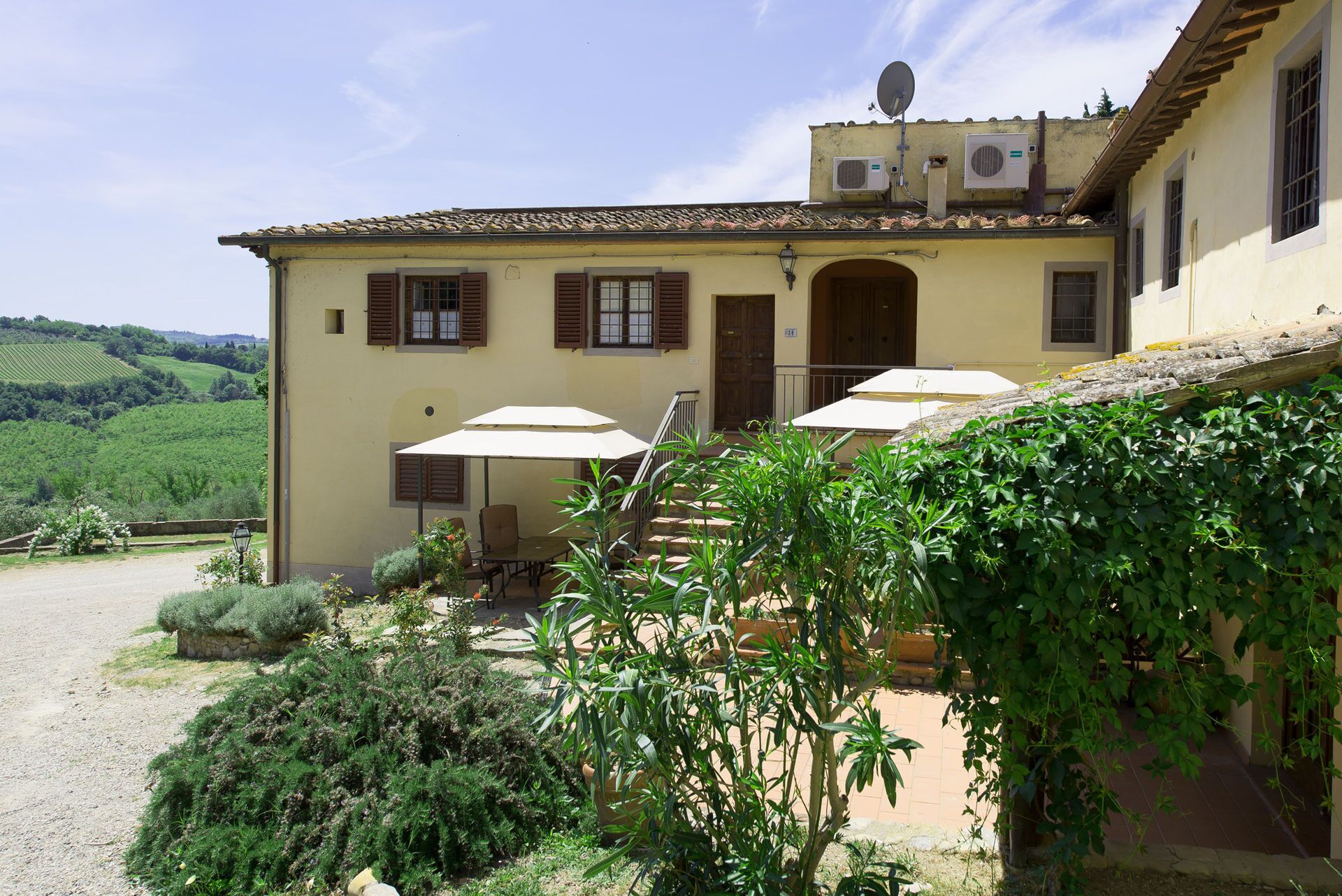 A building with a yellow facade, a tiled roof, and a staircase leading to a door. There are two outdoor umbrellas and a table with chairs.