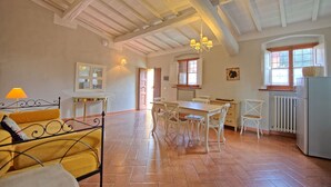 A spacious kitchen with a dining area, a bed with a yellow headboard, and a large window providing natural light. - Montegufoni 4  - Apartment with swimming pool (Montegufoni)