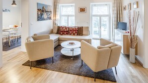 A modern living room with a sofa, two armchairs, a coffee table, and a TV on a stand. There are large windows with curtains, a dining area in the background, and a chandelier. - ❤️ Cozy, Spacious, Quiet, Great Central Location (Praha 3)