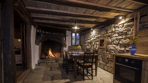 A rustic stone fireplace with a fire, wooden dining table with chairs, and a stone wall with a window.