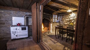 A rustic stone-walled room with a fireplace, wooden ceiling beams, a vintage stove, and a dining table with chairs.