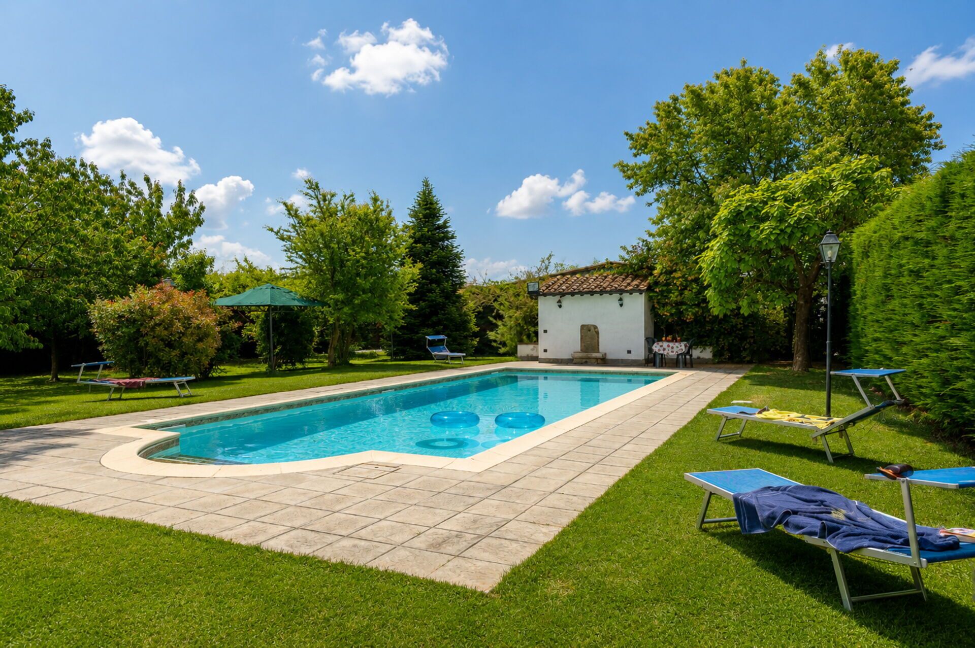 Une piscine rectangulaire, bordée d’une terrasse carrelée, entourée de pelouse et d’arbres. On y trouve des chaises longues et un petit bâtiment avec un toit recouvert de tuiles.