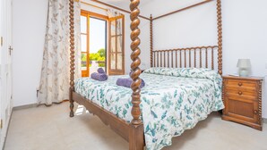 Een hemelbed met een bloemenbedekking, een houten nachtkastje met een lamp, en uitzicht op groen door een open raam.