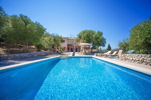 Une piscine rectangulaire à l’eau d’un bleu limpide, entourée de murs en pierre et d’arbres. On aperçoit, au fond, des chaises longues et un espace de repos abrité.