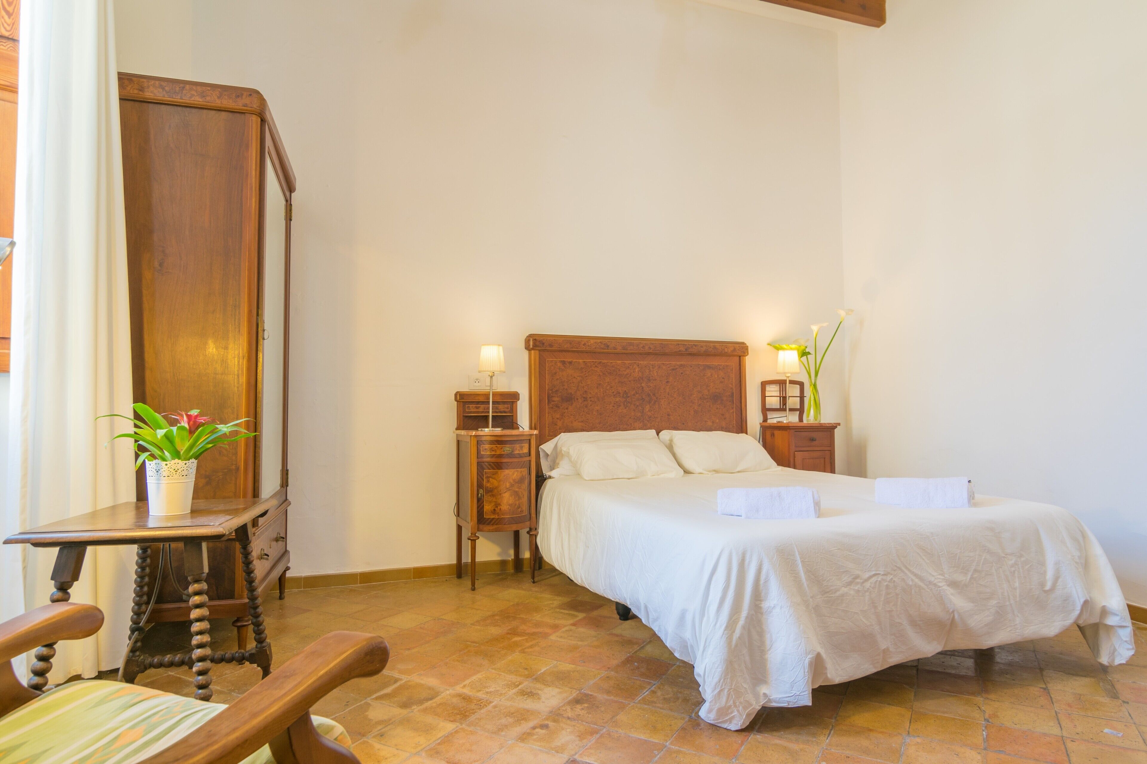 Een slaapkamer met een houten bed, nachtkastje, bijzettafeltje, stoel en een potplant.
