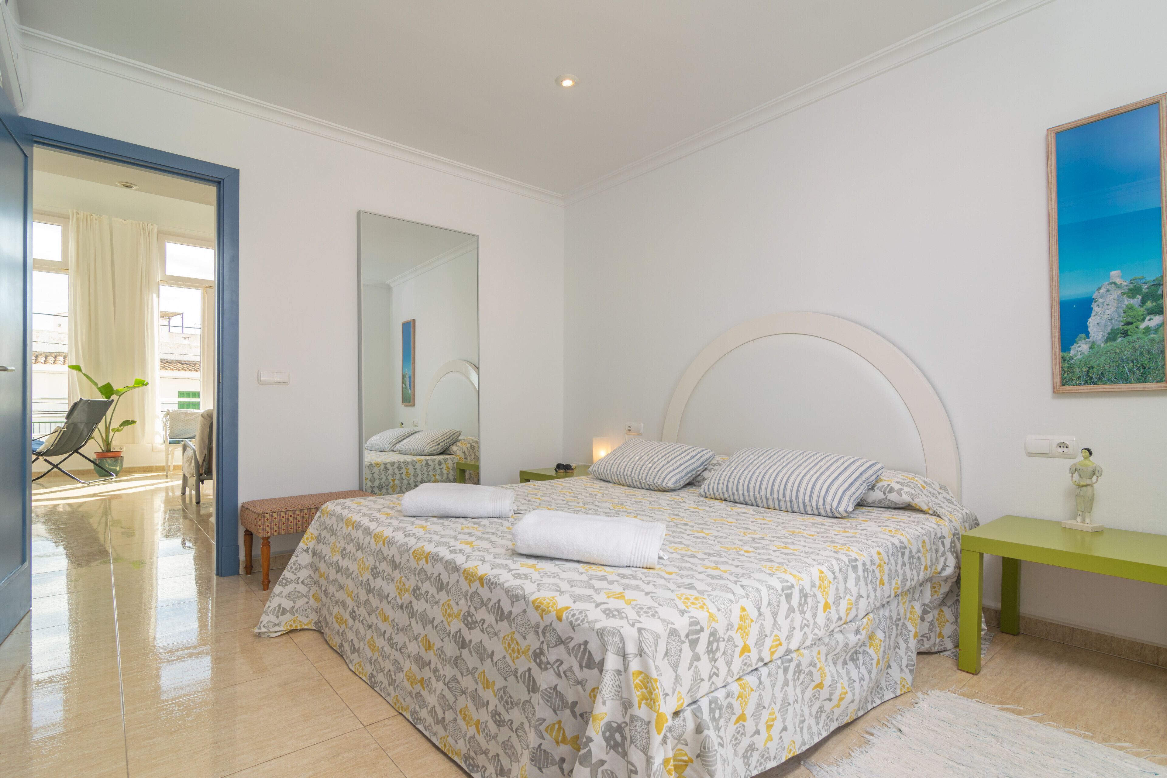 Un dormitorio con cama, mesita de noche, silla, espejo y vistas al exterior.