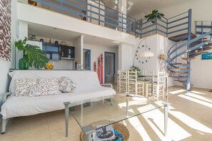 Un amplio salón con una mesa de centro de vidrio, un sofá con cojines estampados y una escalera con barandillas azules.