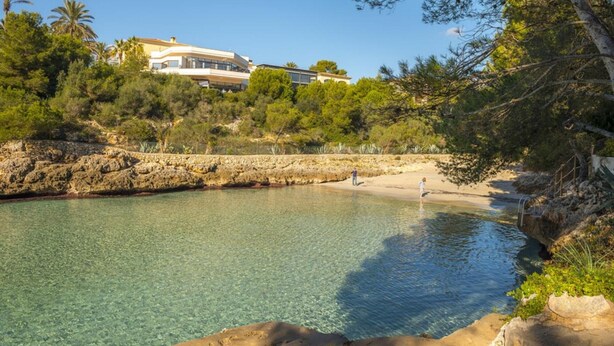 A clear pool surrounded by rocky terrain and dense vegetation, with a large building in the background. - SA CALETA - Villa with private pool in Cala Egos (Santanyi). Free WiFi (Santanyí)