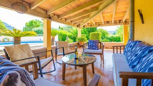 Een terras buiten, met een houten dak, een glazen tafel met een fles en glas, en zitgelegenheid.