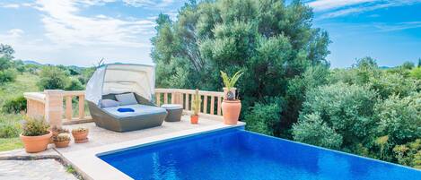 A swimming pool with a lounge area, potted plants, and a view of trees and hills.