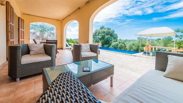 A sunlit patio with wicker furniture, a glass table, and a view of a swimming pool and greenery. - CAN CANALS - Villa with private pool in Artà. Free WiFi. (Artà)
