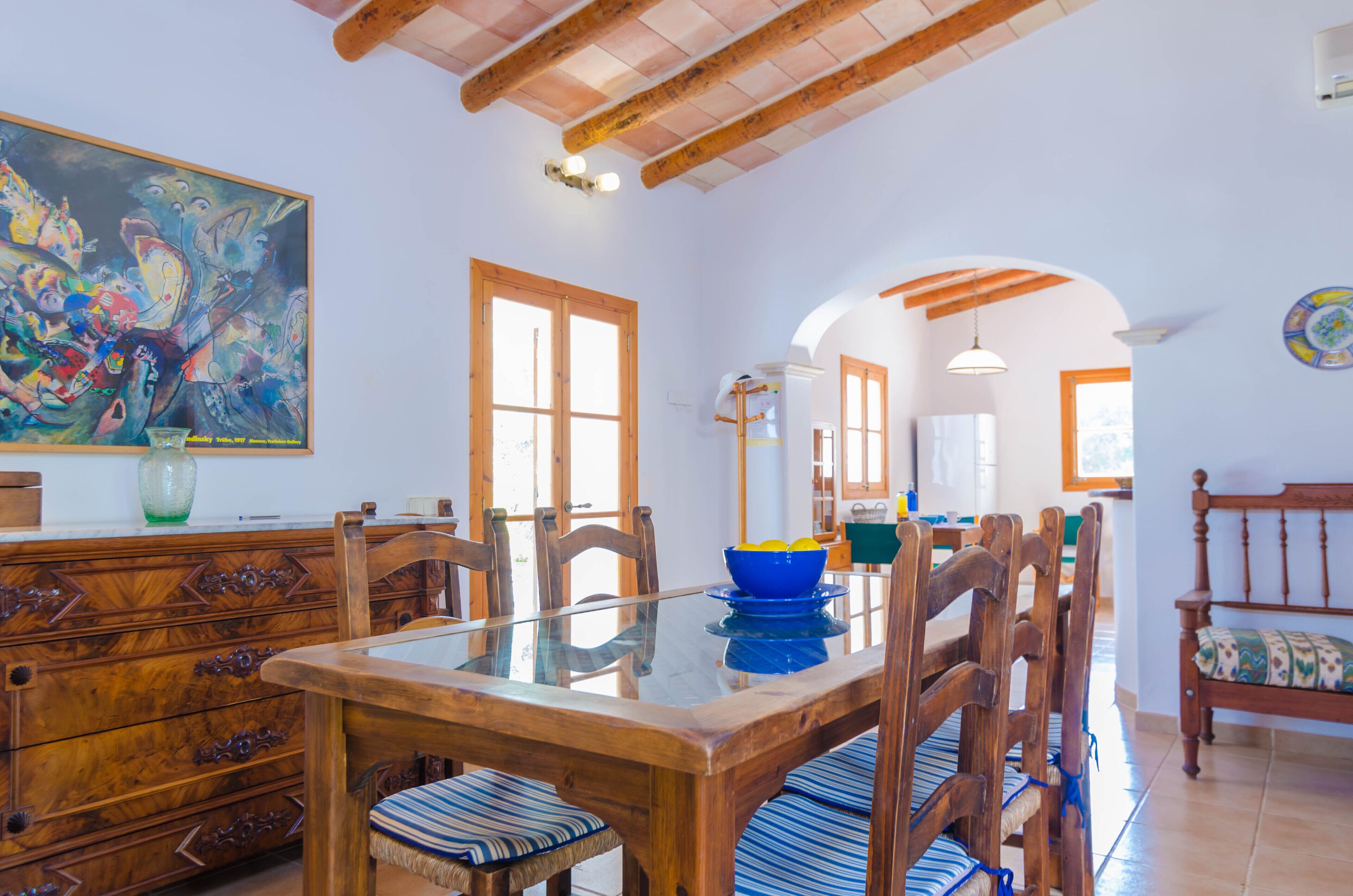Un comedor con una mesa y sillas de madera, un cuadro grande en la pared y vistas a la cocina al fondo.