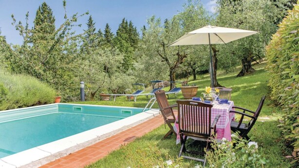 Una piscina con un borde de baldosas, rodeada de césped y árboles. Hay una mesa con mantel a cuadros y sillas, y una sombrilla dando sombra.