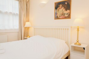 A neatly made bed with white linens, two bedside lamps, a radiator, and a painting on the wall. - Charming Central London Flat (London)
