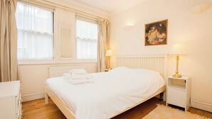 A neatly made bed with white linens, a white nightstand with a lamp, a painting on the wall, and a window with curtains. - Charming Central London Flat (London)