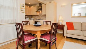 A dining area with a wooden table and chairs, a kitchen in the background, and a sofa with a cushion. - Charming Central London Flat (London)