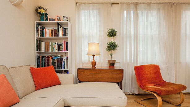 A living room with a beige sectional sofa, a wooden side table with a lamp, a red chair, and a bookshelf filled with books. - Charming Central London Flat (London)