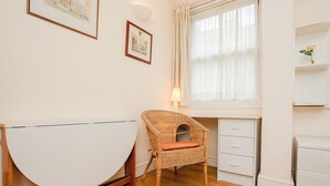 A room with a curved white desk, a wicker chair with a cushion, a lamp, and a window with curtains. - Charming Central London Flat (London)