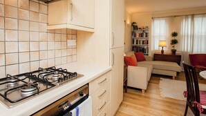 A compact kitchen with a gas stove, adjacent to a living area with a sofa, a bookshelf, and a dining table. - Charming Central London Flat (London)