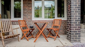 Un espace extérieur aménagé avec deux chaises en bois et une petite table, un mur en briques et des fenêtres donnant sur un quartier résidentiel.