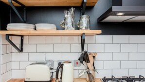 Une cuisine avec une étagère en bois où sont posées des assiettes et des bocaux en verre, un grille-pain, une bouilloire et une cuisinière à gaz.