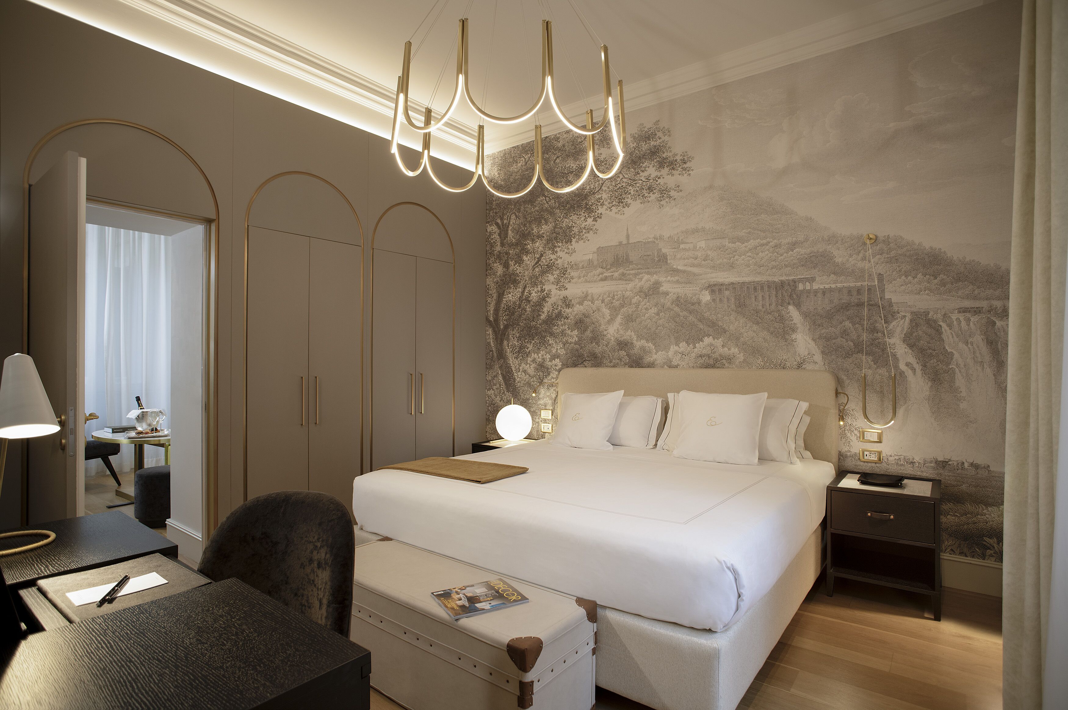 Una habitación de hotel moderna con una cama grande, un escritorio y una silla. Se aprecia una mesita de noche con una lámpara y un mural en la pared.