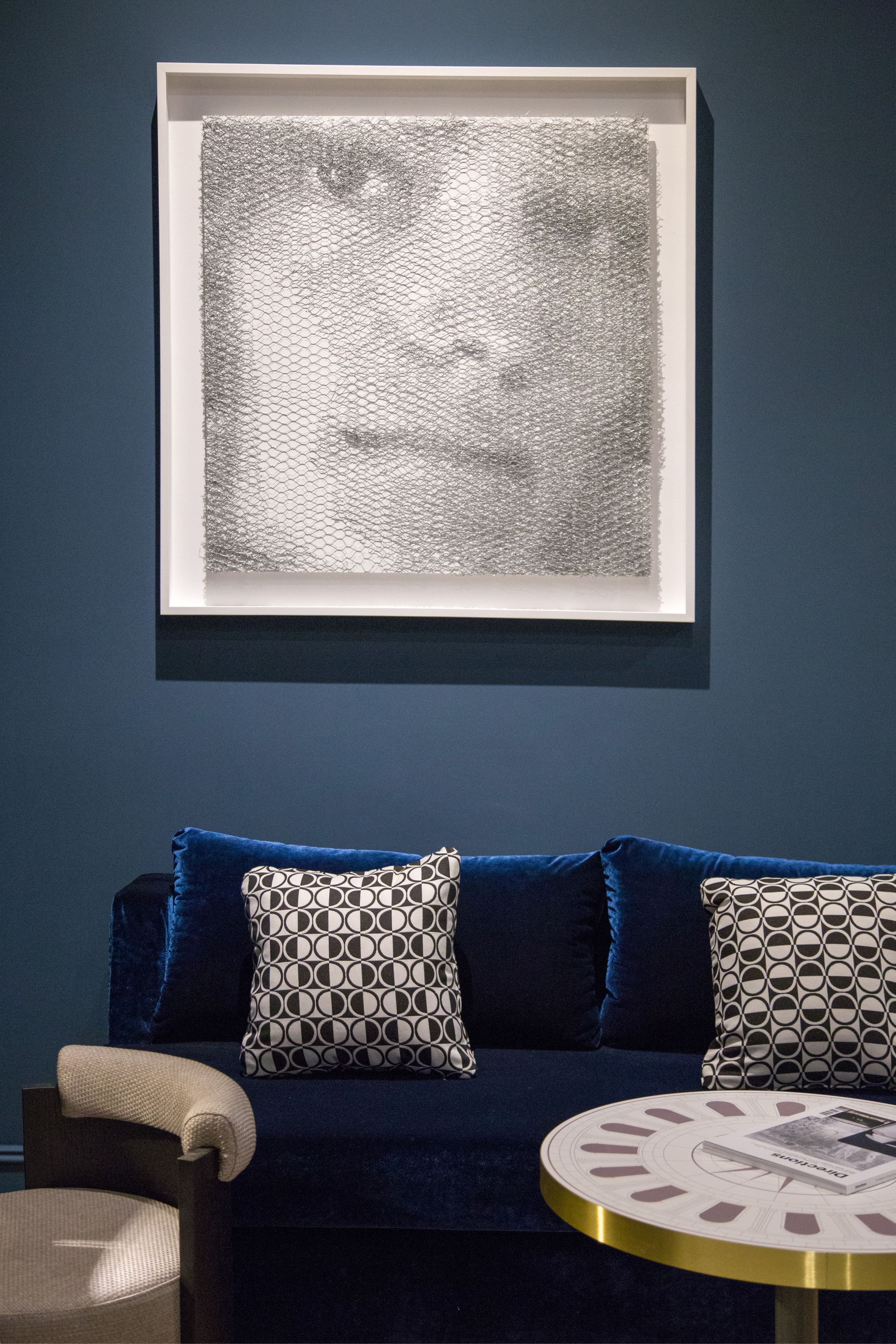 A room with a dark blue sofa, patterned pillows, a framed artwork, and a round table with a book.