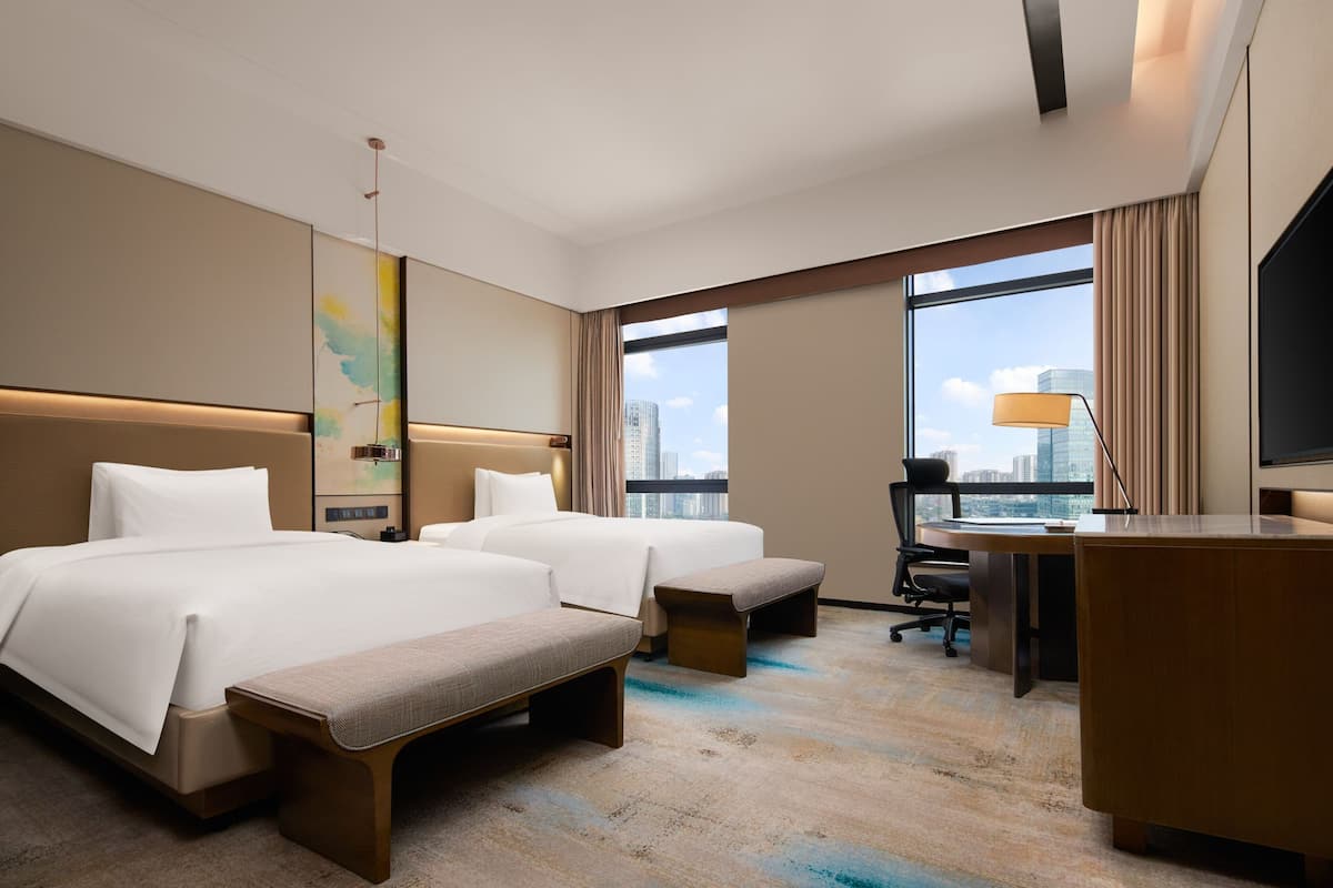 A hotel room with two beds, a desk, a chair, and a large window with a city view.