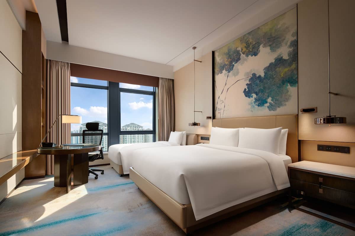 A hotel room with a large bed, a desk, a chair, and a view of the city through the window.