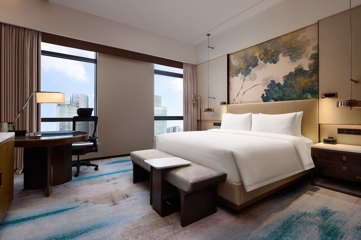 A hotel room with a large bed, a desk, a chair, a bench, and a view of the cityscape through large windows.