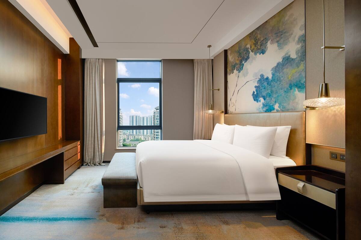 A modern hotel room with a large bed, a flat-screen TV, a bench, and a city view.