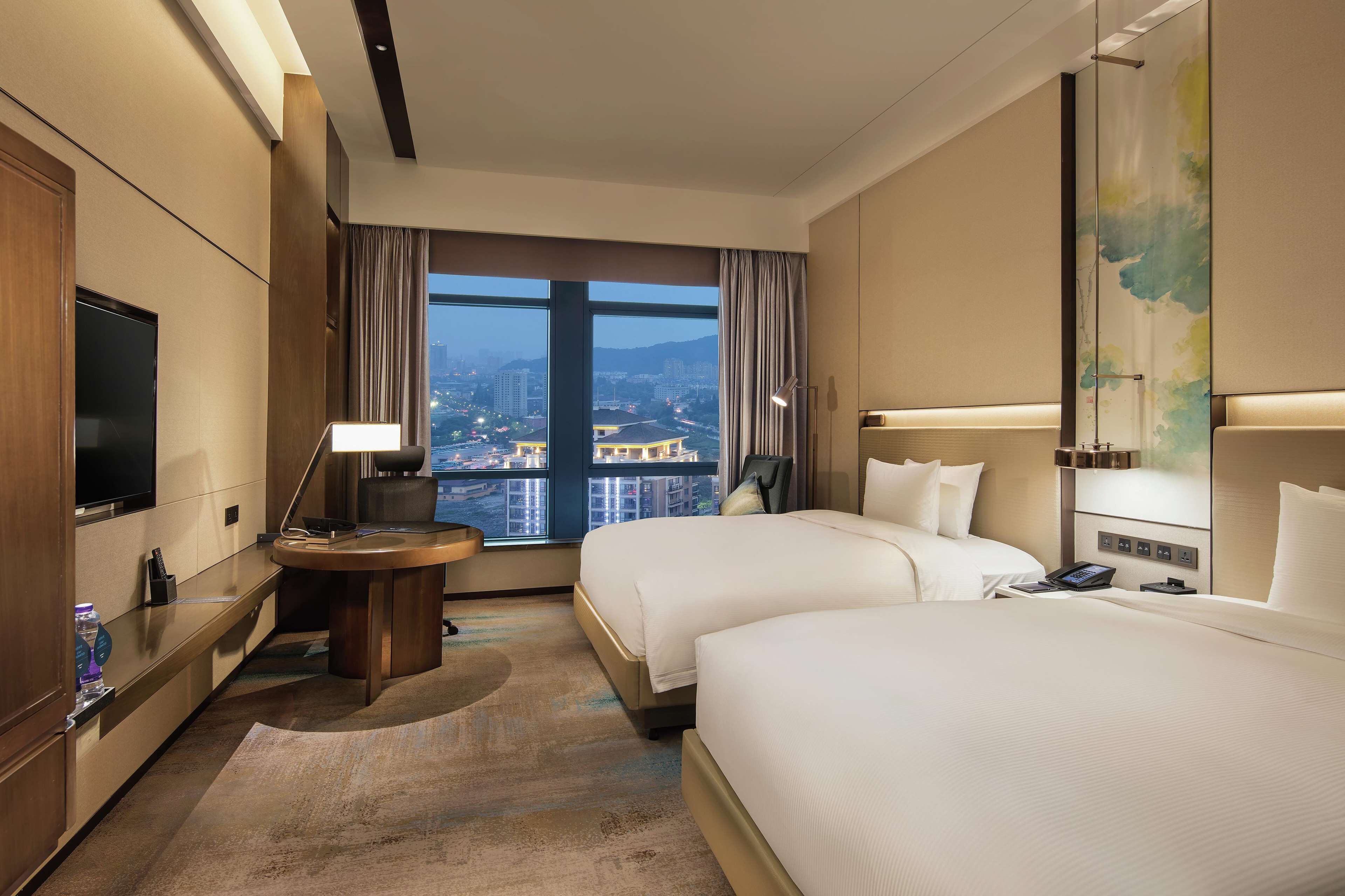 A hotel room with two beds, a TV, a desk with a lamp, and a view of the cityscape.