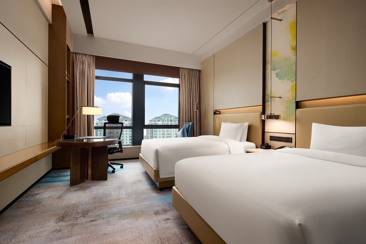 A hotel room with two beds, a desk, a chair, a TV, and a large window with a city view.