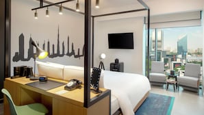 Moderni hotellihuone, jonka seinällä on kaupungin siluettia esittävä suuri seinämaalaus. Huoneessa on työpöytä lampun kanssa, sänky, televisio ja parveke, jolta avautuu näkymä kaupunkiin.