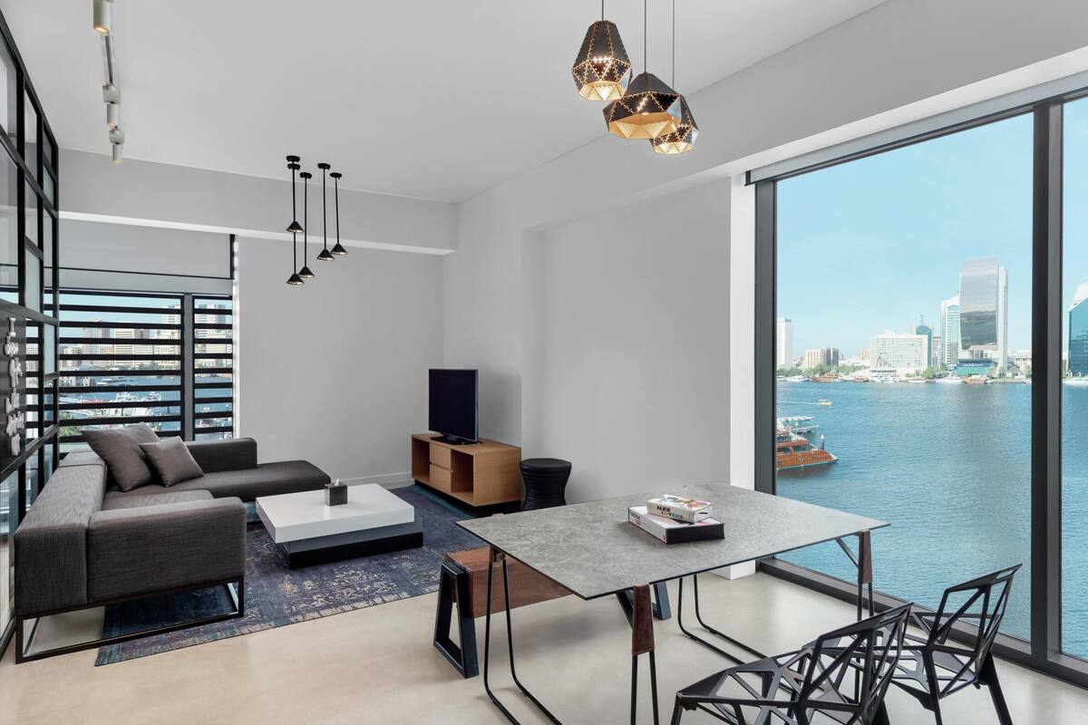 A modern living room with a sofa, coffee table, and dining area, overlooking a body of water with buildings in the distance.