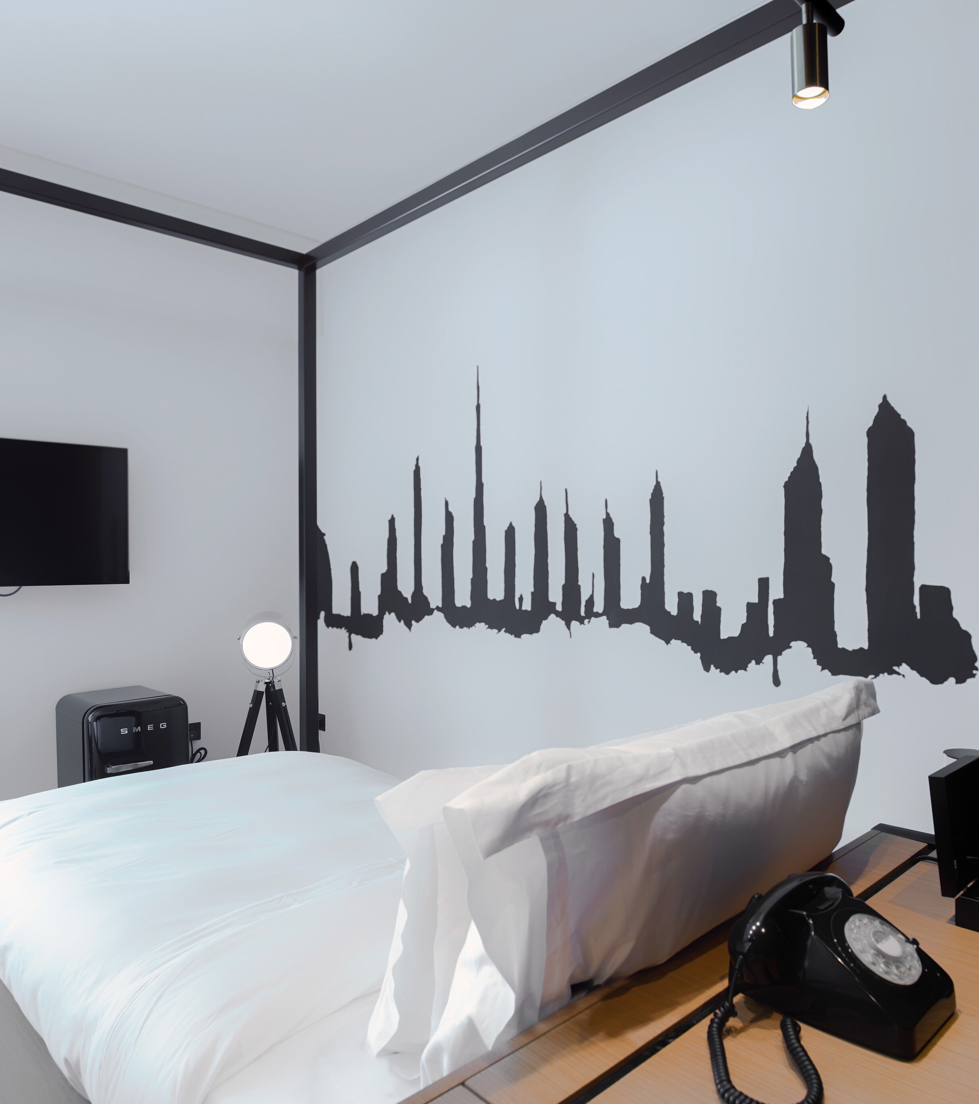 A hotel room with a bed, a desk, a lamp, a phone, and a wall decal of a city skyline.