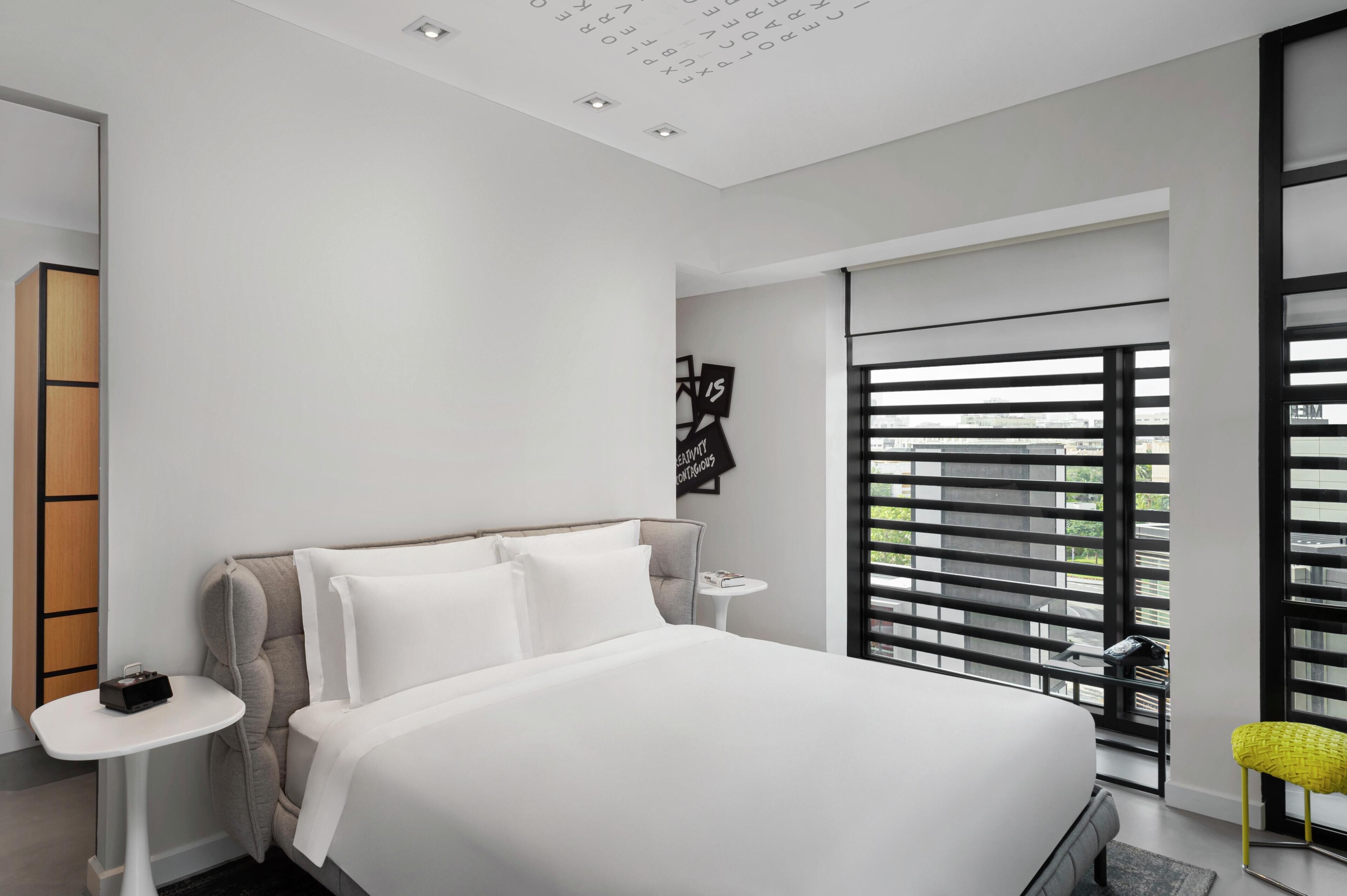 A modern hotel room with a large bed, a small table, a chair, and a view through floor-to-ceiling windows.