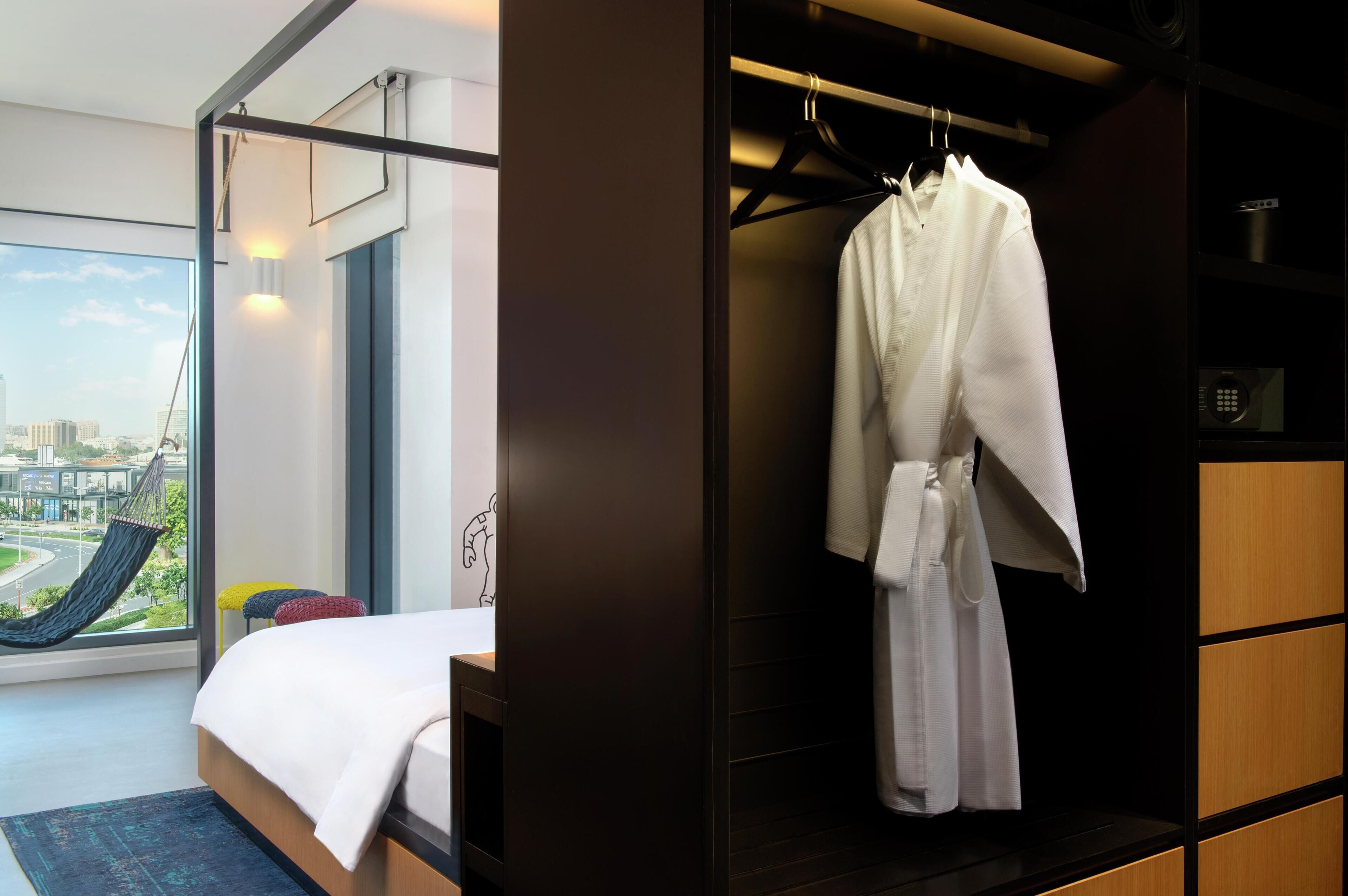 A modern hotel room with a large bed, a view of a cityscape, a hanging chair, and a walk-in closet with a robe hanging.